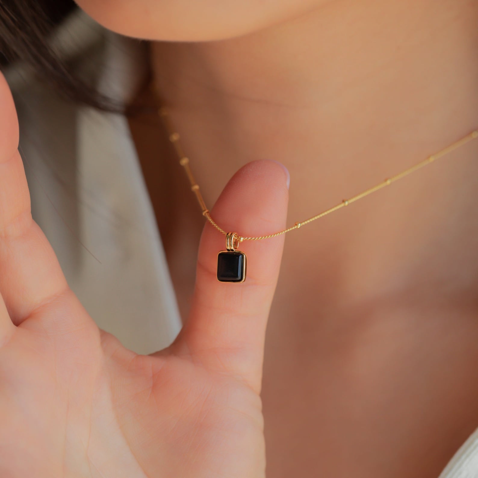A hand holds the Waverly Black Necklace, a gold satellite chain with a small black enamel pendant, near a person's neck.