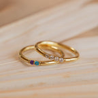 A Mini Birthstone Ring in Rose Gold, featuring three colored gemstones, and another gold ring with four clear stones rest elegantly on a wooden surface.