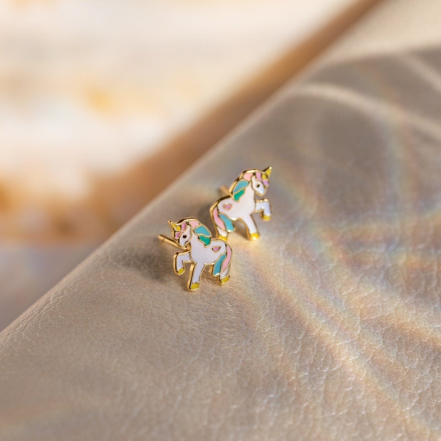 Unicorn Studs: Gold-outlined small unicorn earrings with pastel enamel details rest on a shimmery beige surface—an adorable gift for girls.