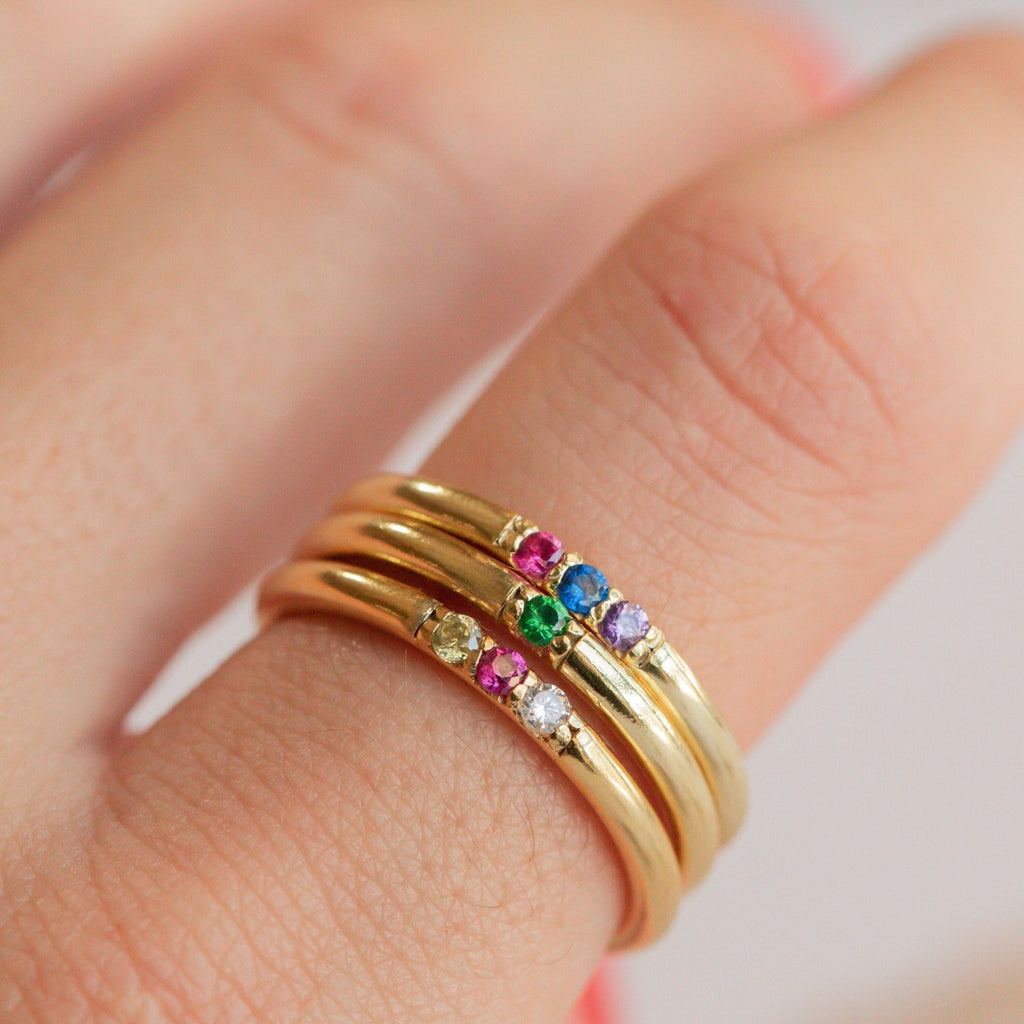 Three gold rings, including the Mini Birthstone Ring adorned with colorful gemstones, are stacked on a person's finger against a blurred background.
