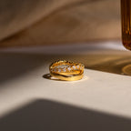 The Wave Diamond Duo Ring, featuring embedded gemstones, rests on a white surface illuminated by soft, warm light.
