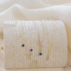 Five dainty Custom Birthstone Necklaces with colored gemstone pendants are displayed on a textured, curved off-white surface—perfect as meaningful jewelry or the ultimate personalized gift.