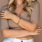 A woman with wavy blonde hair, arms crossed, wears the Aris Hinge Bangle, a beige ribbed top, and white jeans.