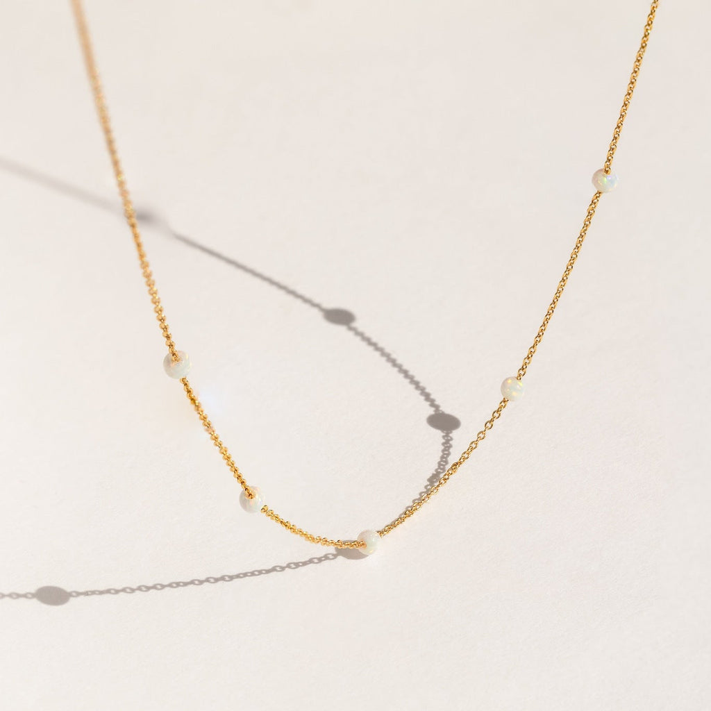 Delicate gold chain necklace with evenly spaced small opal beads, shown casting soft shadows on a light cream background.