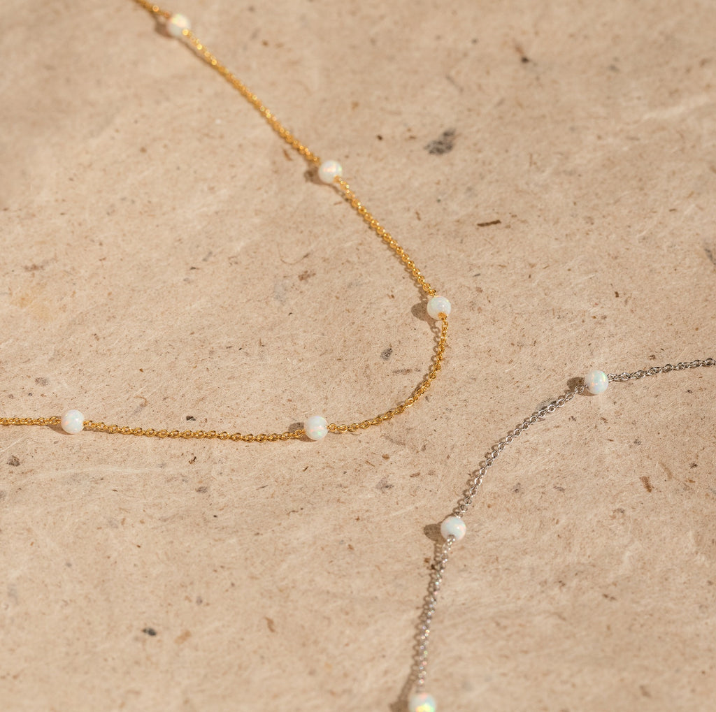 The Opal Station Necklace features gold and silver chains with small white opals in a minimalist design, showcased on a beige textured surface.