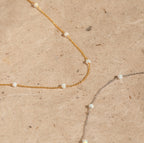 The Opal Station Necklace features gold and silver chains with small white opals in a minimalist design, showcased on a beige textured surface.