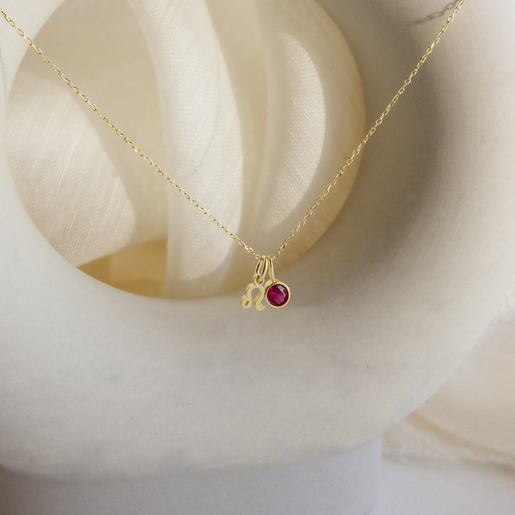 The Zodiac Birthstone Necklace features a Leo charm and a small red gemstone pendant, displayed on a white curved surface—an elegant, personalized jewelry piece ideal for those who love unique birthstone accessories.