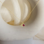 The Zodiac Birthstone Necklace features a Leo charm and a small red gemstone pendant, displayed on a white curved surface—an elegant, personalized jewelry piece ideal for those who love unique birthstone accessories.