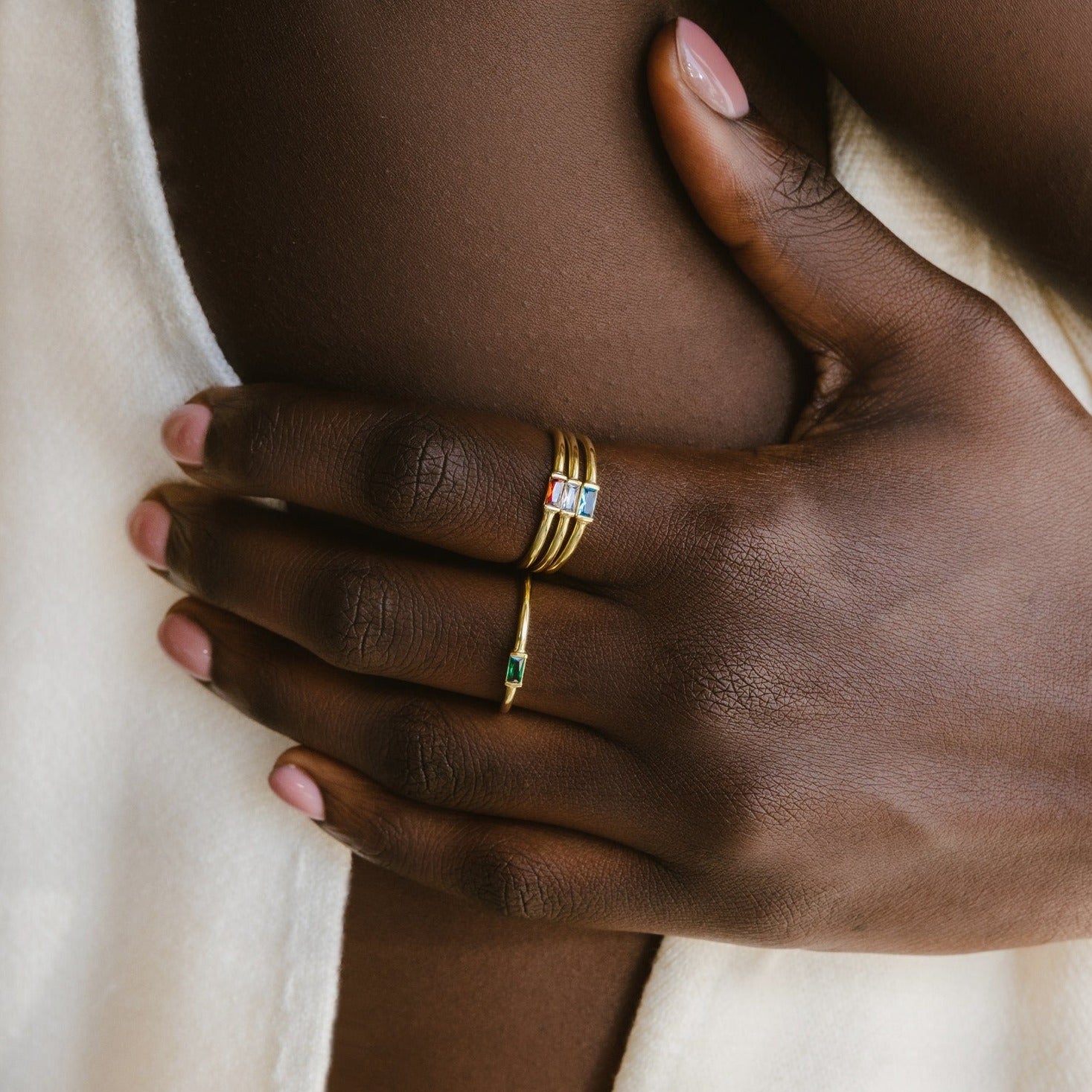 Close-up of a hand with gold stacking rings, including the Baguette Birthstone Ring adorned with colorful gems, resting on a bare shoulder.