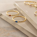 The Marquise Birthstone Ring and two gold dainty rings with colorful gemstones are displayed on reflective glass surfaces.