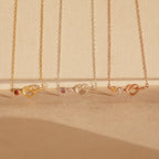 Four delicate Love Knot Birthstone Necklaces in gold, silver, and rose gold with small gemstones, perfect for celebrating family, displayed on a beige background.