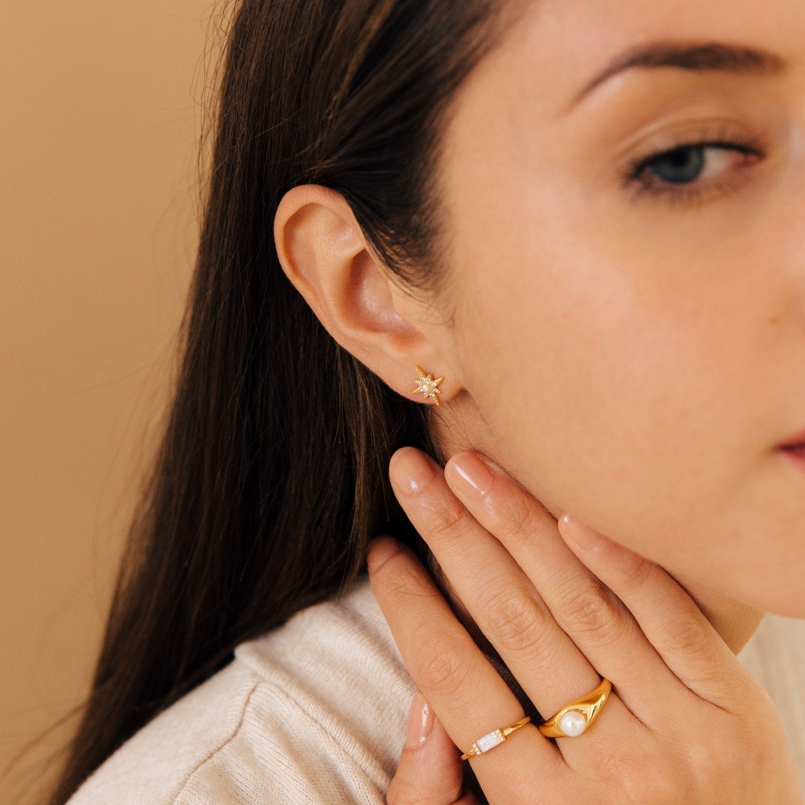 Woman with long brown hair wears gold Starburst Studs and two gold rings as she touches her neck.