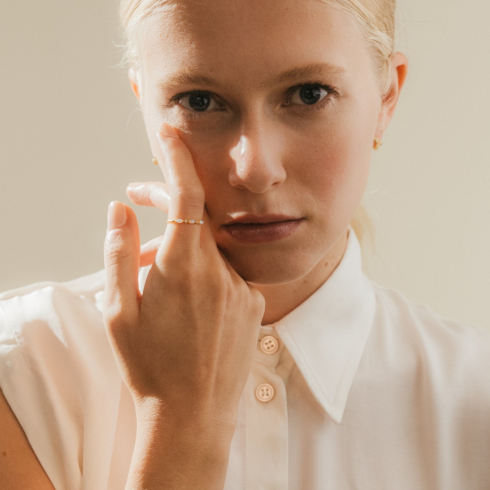 A woman in a white button-up shirt touches her face, wearing the Alessia Trio Opal Ring and stud earrings.