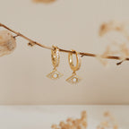 The Aleah Opal Evil Eye Huggies feature eye-shaped pearl and crystal charms, capturing the allure of evil eye jewelry and elegantly displayed against a beige background.