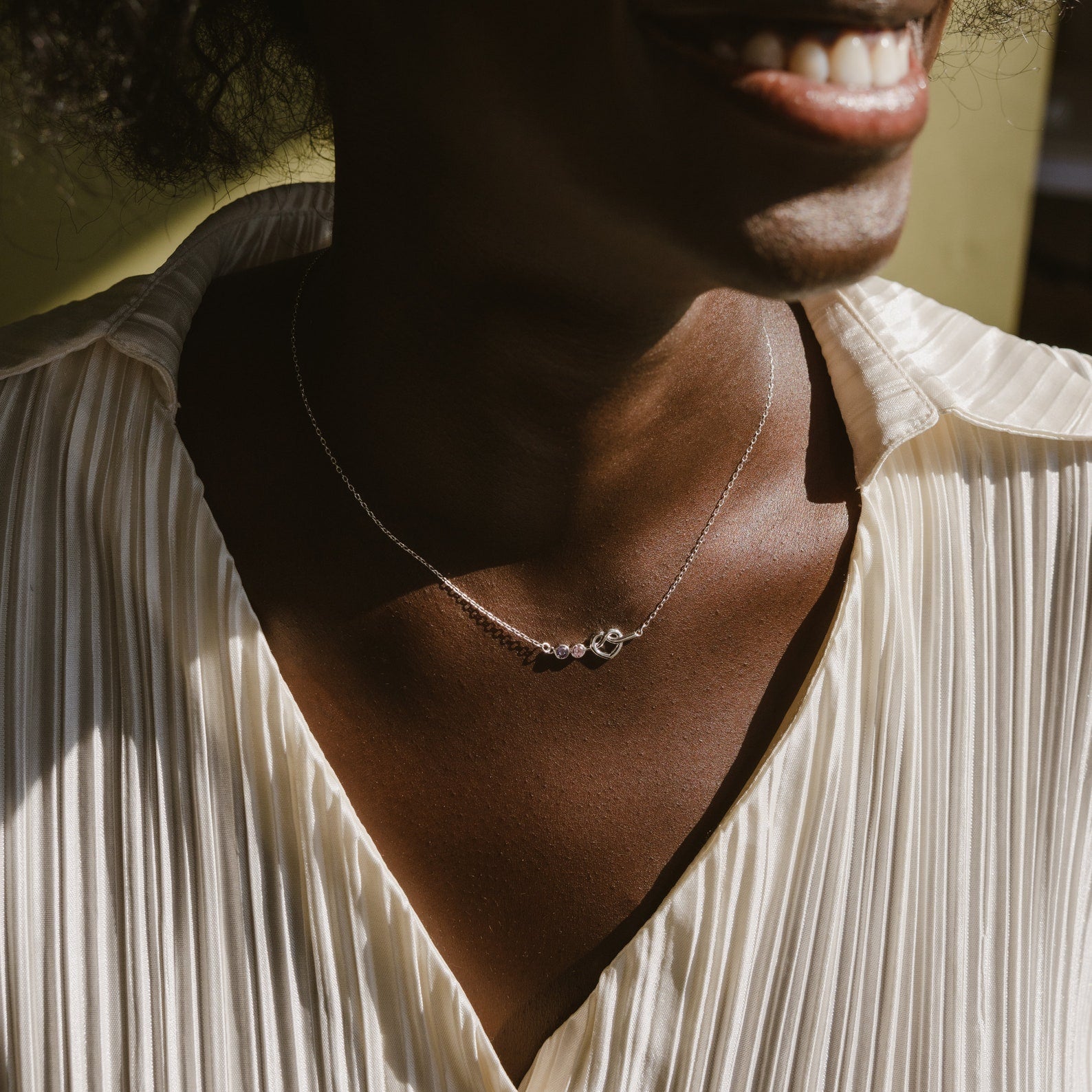 Smiling woman wearing a pleated white blouse and a delicate Love Knot Birthstone Necklace, adorned with small personalized pendants.