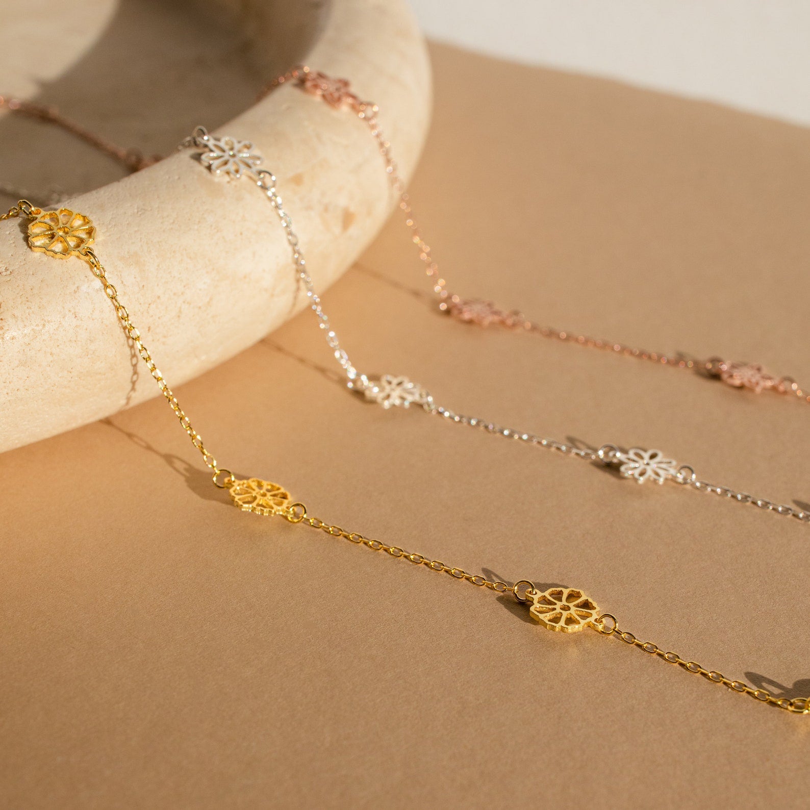 The Flower Station Necklace features a delicate gold, silver, and rose gold chain with a floral charm—an elegant floral jewelry piece and perfect gift for her, beautifully displayed on a beige surface.