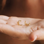 A hand holds two Eclipse Moon Studs—gold crescent moon earrings adorned with pearls and tiny beads.