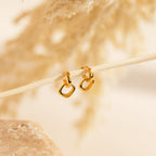 Reagan Duo Huggies dangle elegantly from a slender branch, featuring gold geometric hoops that shine against a subtle, neutral backdrop.