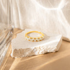 The Opal Drop Eternity Ring, a vintage-inspired gold piece with shimmering white gems, is displayed on a rough white stone in soft natural light against a neutral background.