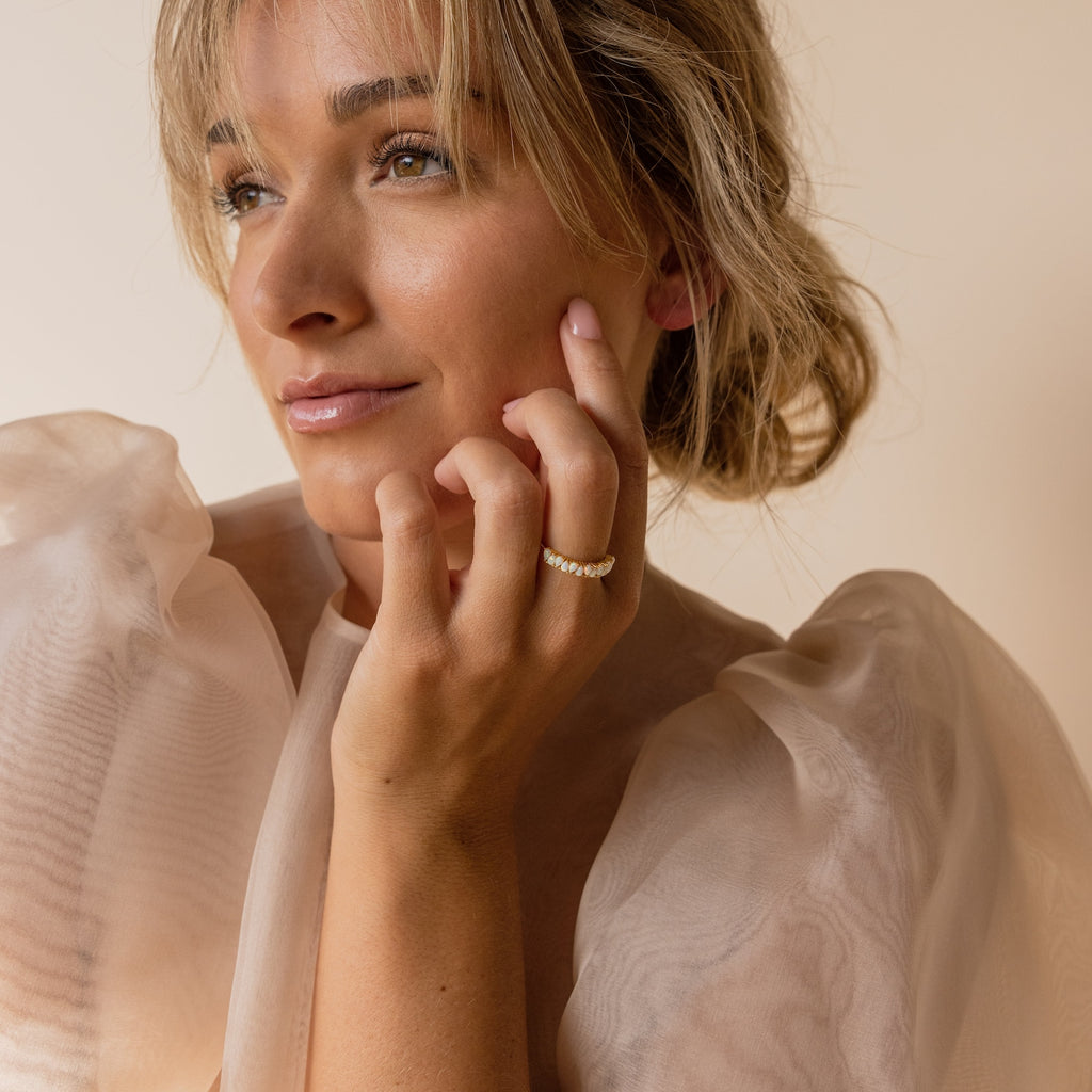 A woman with a gentle smile wears a sheer, puffy-sleeved top, showcasing the Opal Drop Eternity Ring—a vintage-inspired gemstone piece—on her finger.