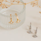 Aleah Opal Evil Eye Huggies—gold and silver huggie hoop earrings with opal gemstone evil eye details, displayed on a glass dish and cream surface. A stunning piece of evil eye jewelry.