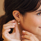 Woman with brown hair smiles softly, gently touching her ear adorned with Aleah Opal Evil Eye Huggies.
