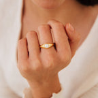 A hand showcases the Penelope Pearl Signet Ring, featuring a central freshwater pearl in a gold setting, set against a soft beige fabric background.