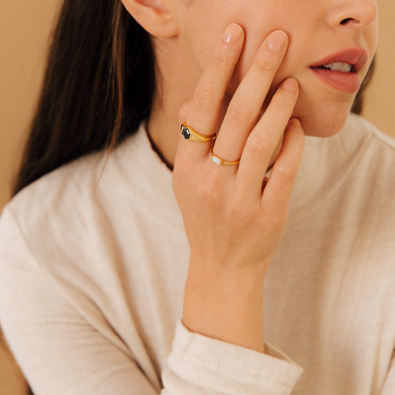 A woman with long hair touches her face, wearing the Ember Signet Ring and gold rings, styled in a cream-colored top against a beige background.
