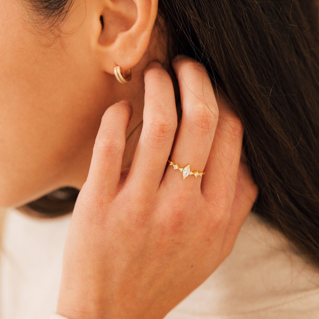 A woman touches her hair, wearing the Harlow Diamond Ring in 18K Gold and a small gold hoop earring.