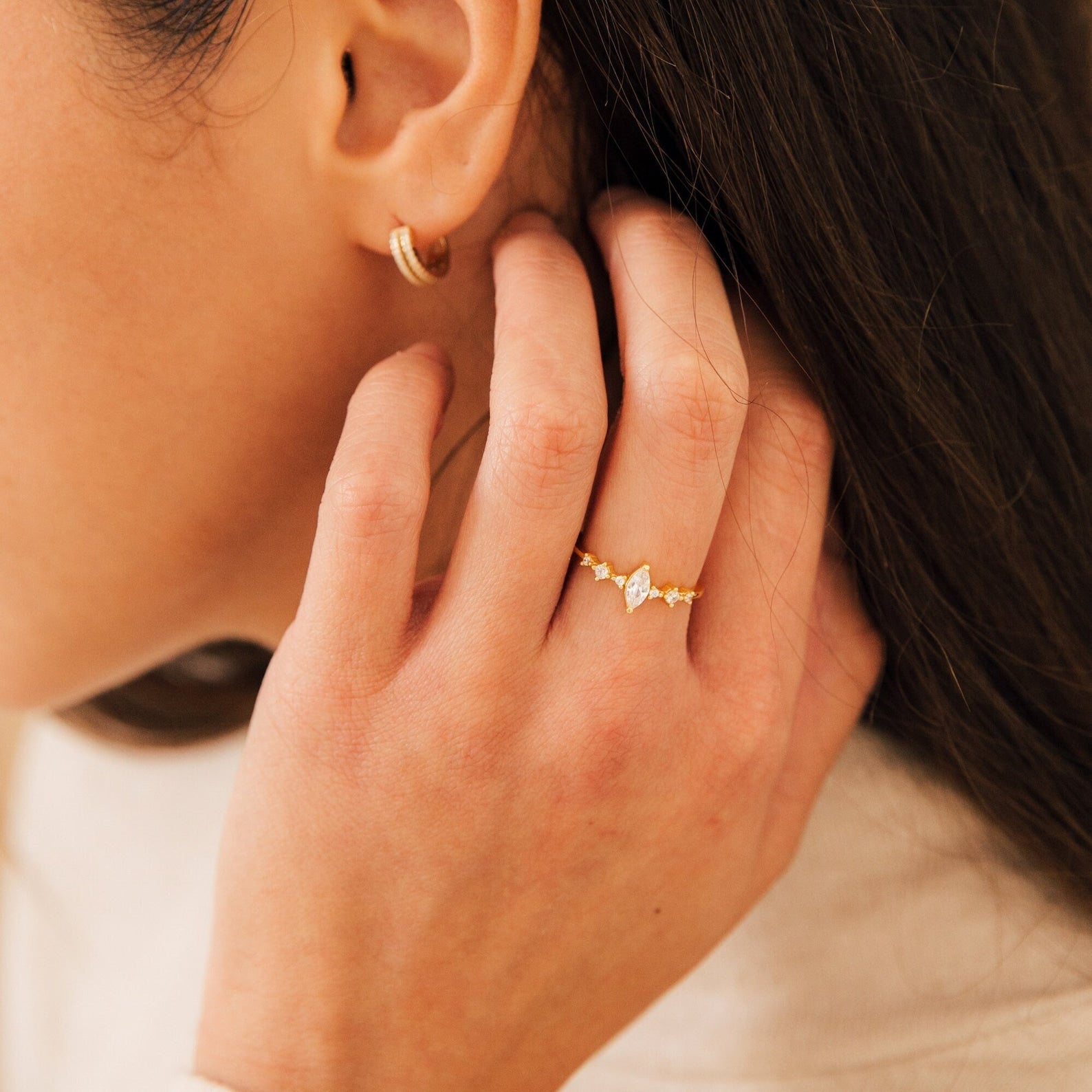 A woman touches her hair, wearing the Harlow Diamond Ring in 18K Gold and a small gold hoop earring.
