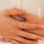 Close-up of hands wearing the Mosaic Wave Eternity Ring, a delicate gold band adorned with small white stones, shown against a cream fabric background.