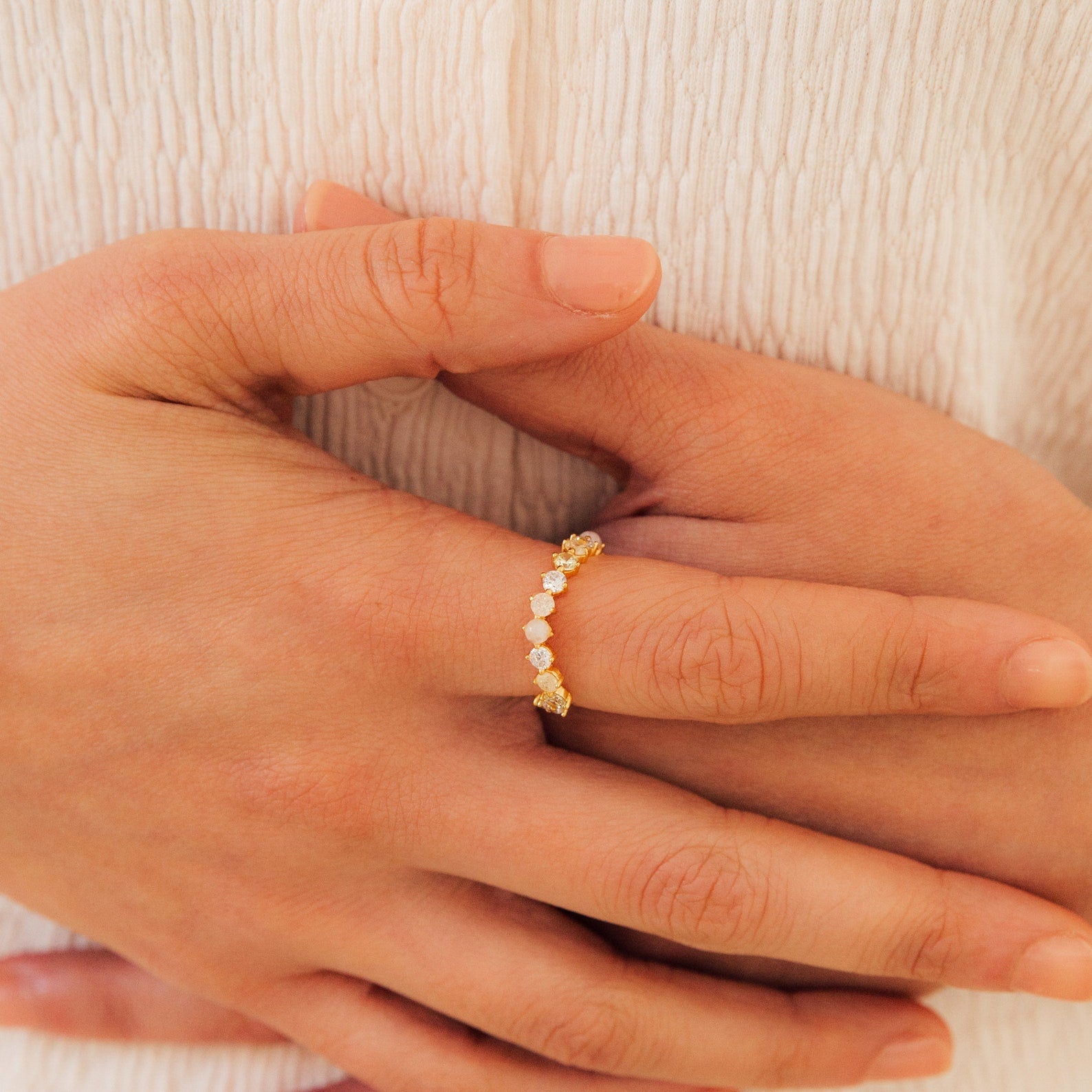 Close-up of hands wearing the Mosaic Wave Eternity Ring, a delicate gold band adorned with small white stones, shown against a cream fabric background.
