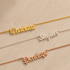 Three Old English Name Necklaces in curb chains lay on a beige surface, personalized with "Charlotte," "Skylar," and "Penelope" in different metals, each featuring a custom charm for a modern look.
