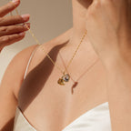 A woman in a white satin top holds the Initial Heart Locket Necklace, a gold heart pendant with two charms, adding a personalized jewelry touch near her neck.