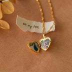 The Initial Heart Locket Necklace, a gold heart-shaped locket with engraved initial and photos inside, rests beside a note reading "Oh my love" on a brown surface.
