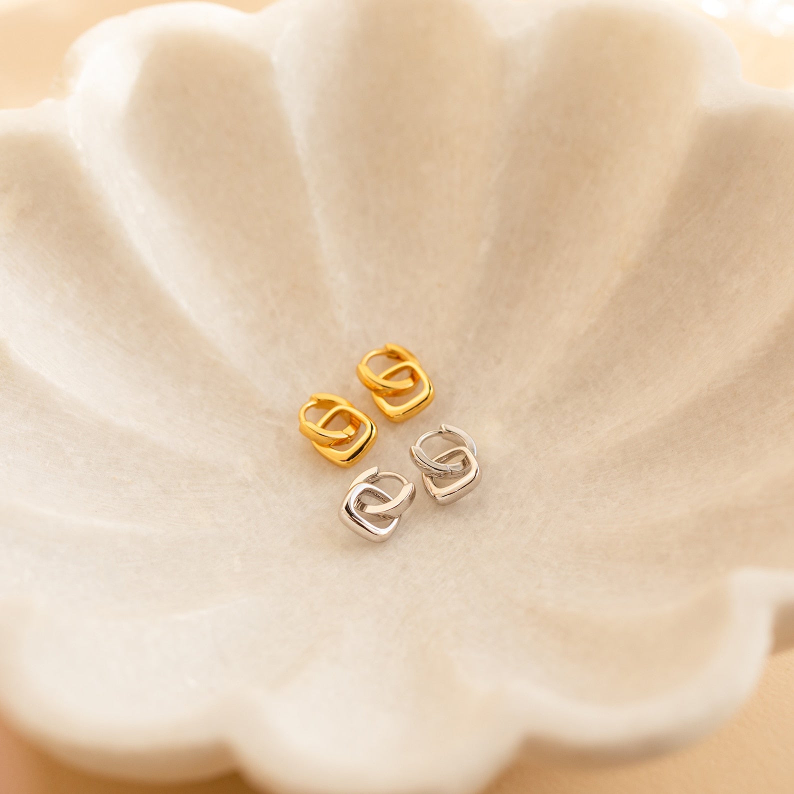 Two pairs of chic geometric gold and silver Reagan Duo Huggies, including minimalist hoops, displayed on a scalloped cream-colored dish.