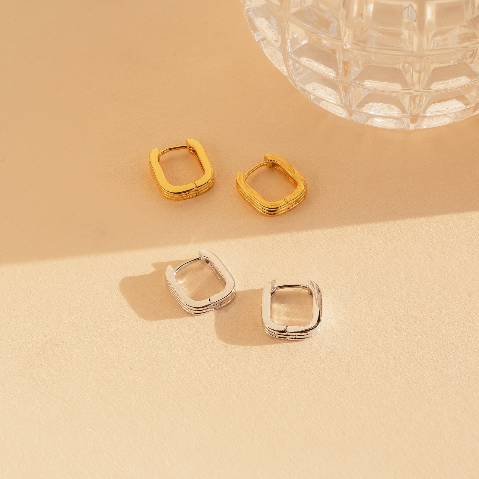 Two pairs of Ribbed Link Hoops—one gold, one silver—rest on a beige surface beside a glass object. Each showcases modern elegance with their geometric, ribbed rectangle design for a chic, contemporary vibe.