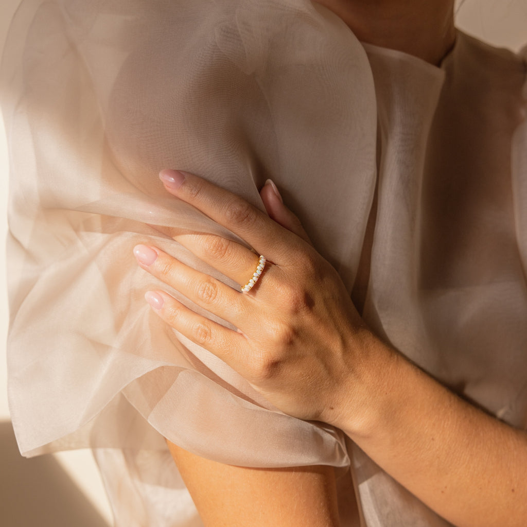 A woman in a sheer, light dress with her hand on her shoulder wears a vintage-inspired Opal Drop Eternity Ring, embellished with small gemstones.