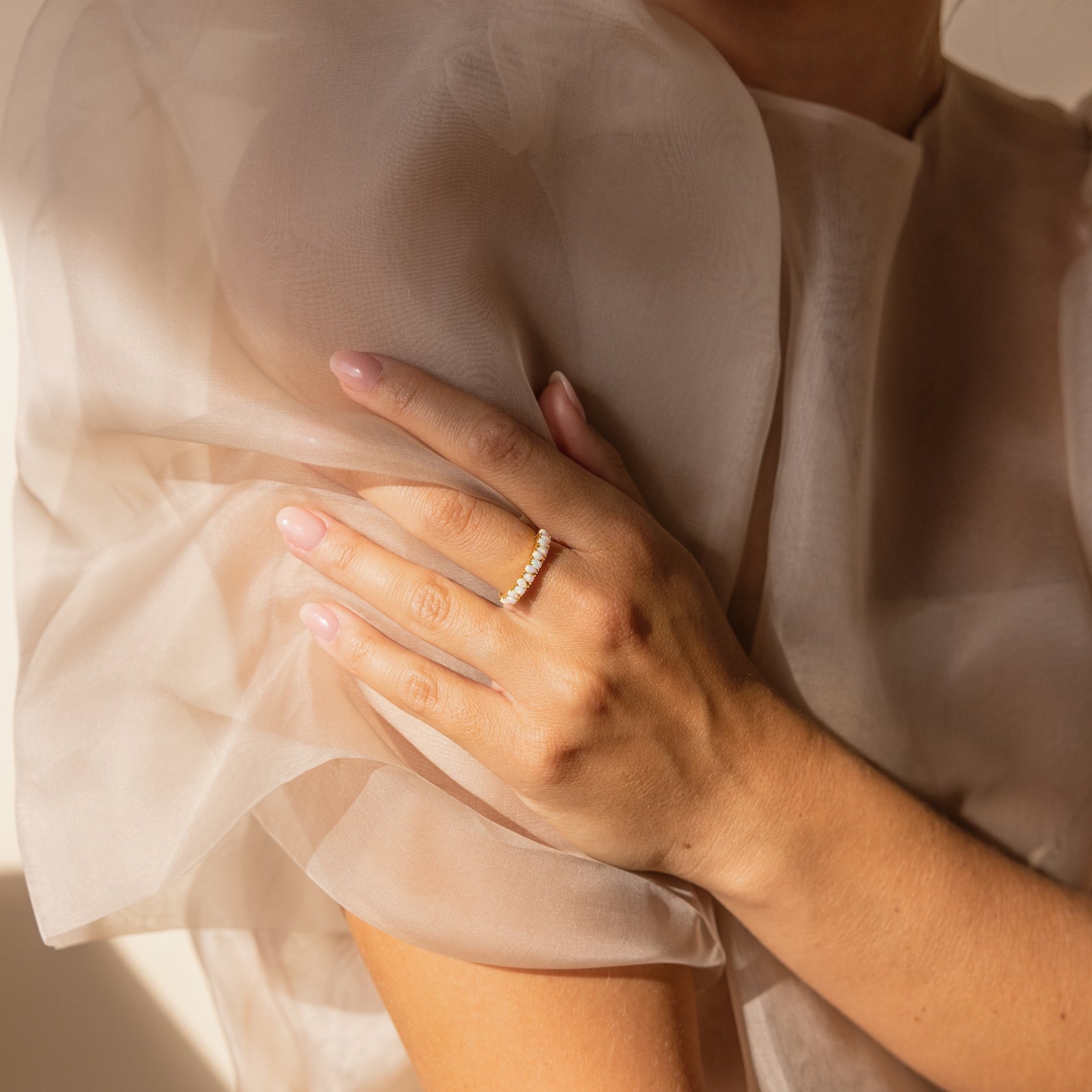 A woman in a sheer, light dress with her hand on her shoulder wears a vintage-inspired Opal Drop Eternity Ring, embellished with small gemstones.