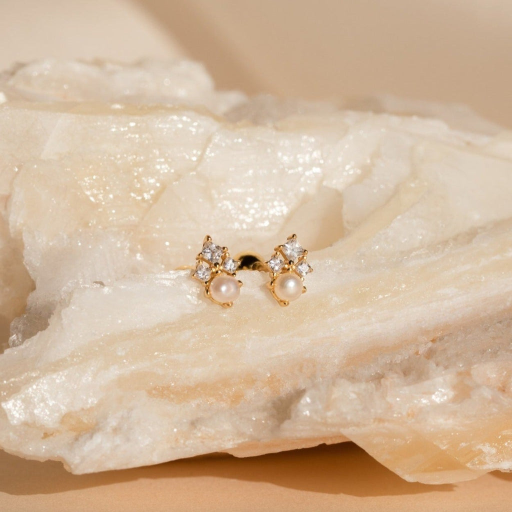 Angelina Pearl Studs, featuring elegant pearls and sparkling crystals, are displayed on a textured, light-colored stone background.