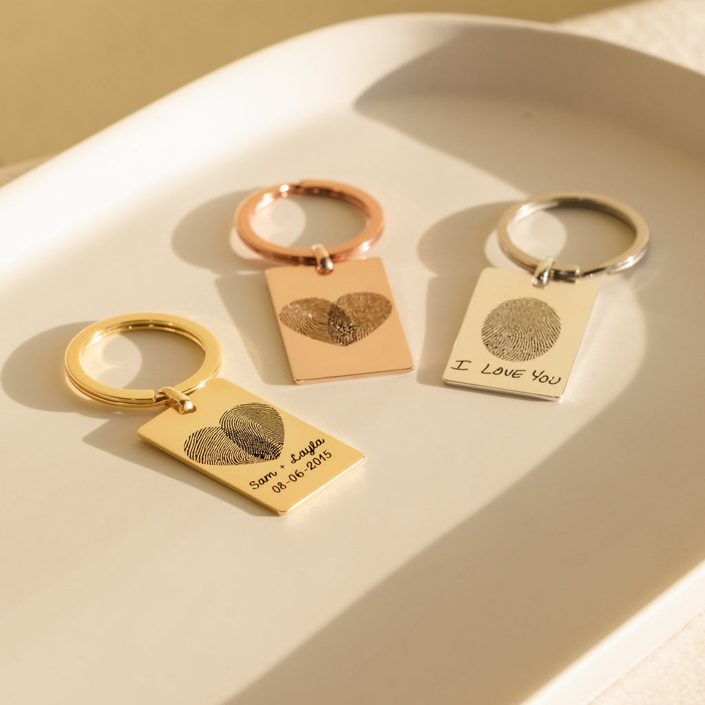 Three Fingerprint Rectangle Keychains with engraved fingerprints forming hearts rest on a white tray in sunlight—a perfect personalized keepsake.