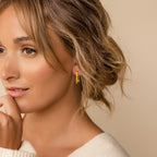 Woman with wavy hair wearing minimalist Large Ribbed Link Hoops and a cream sweater, smiling softly against a beige background.