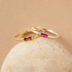 Two delicate Baguette Birthstone Rings with rectangular gemstones—one gold with orange, one silver with pink—rest on a smooth rock. Personalize this ring for a unique way to celebrate your story or as a thoughtful birthstone gift.