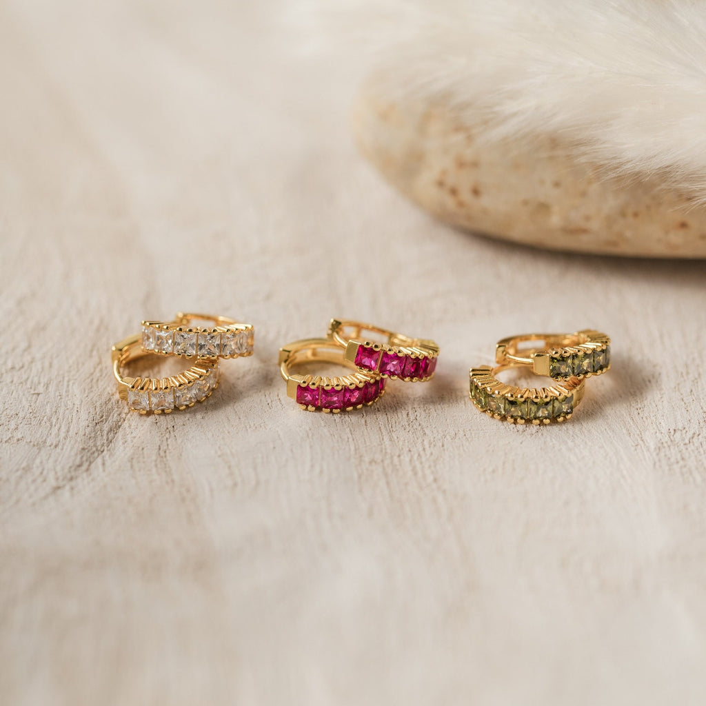 A set of three pairs of Genevieve Birthstone Huggie earrings in gold, featuring sparkling square-cut gemstones in clear diamond, vibrant ruby red, and peridot, displayed on a neutral textured surface with a soft beige stone accent.