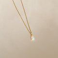 A gold chain necklace featuring a small, iridescent teardrop charm pendant—the Tabitha Opal Drop Necklace—displayed on a light beige background, perfect as a romantic gift.