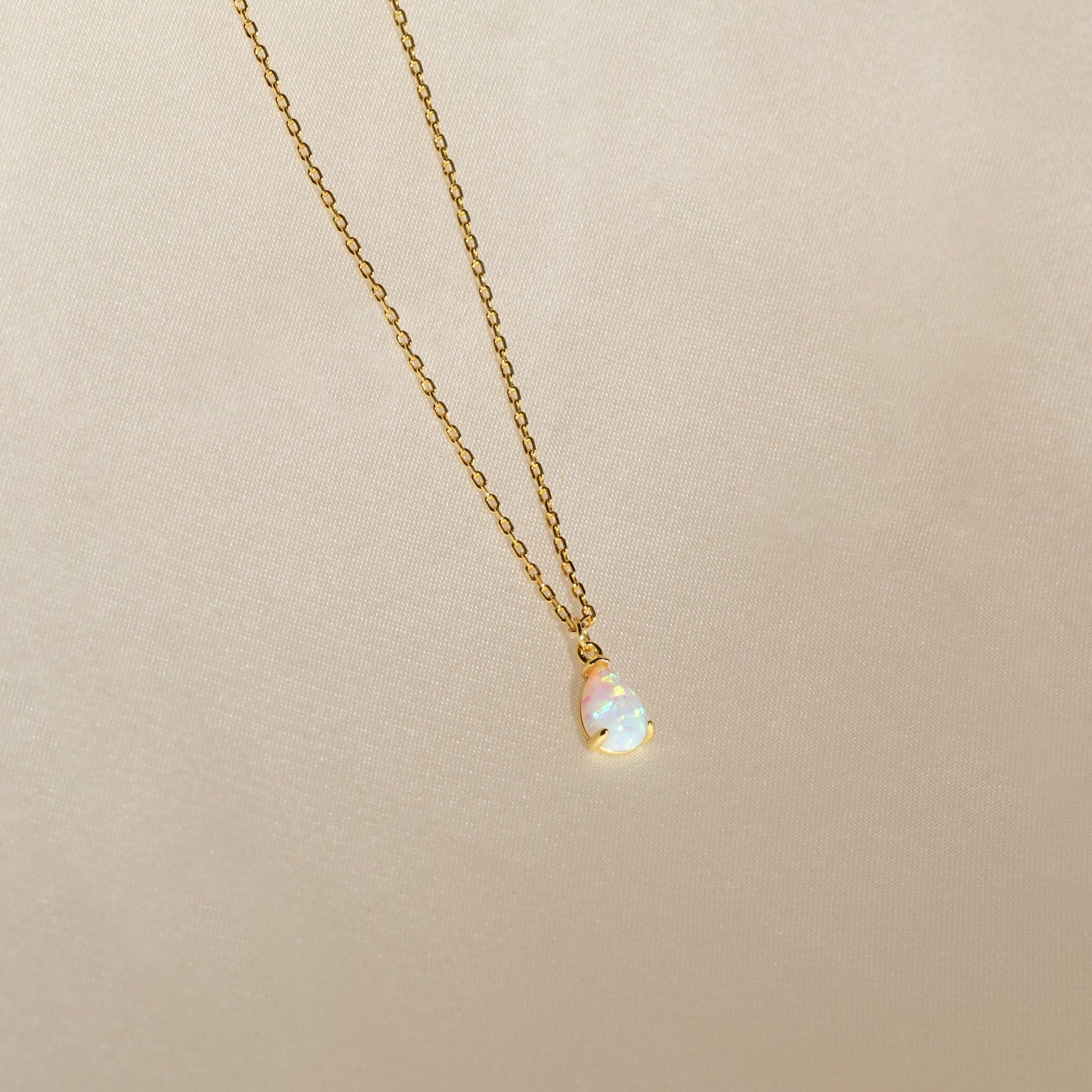 A gold chain necklace featuring a small, iridescent teardrop charm pendant—the Tabitha Opal Drop Necklace—displayed on a light beige background, perfect as a romantic gift.