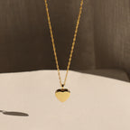 The Initial Heart Locket Necklace, a gold heart-shaped pendant with an engraved initial, is displayed against a soft neutral background.