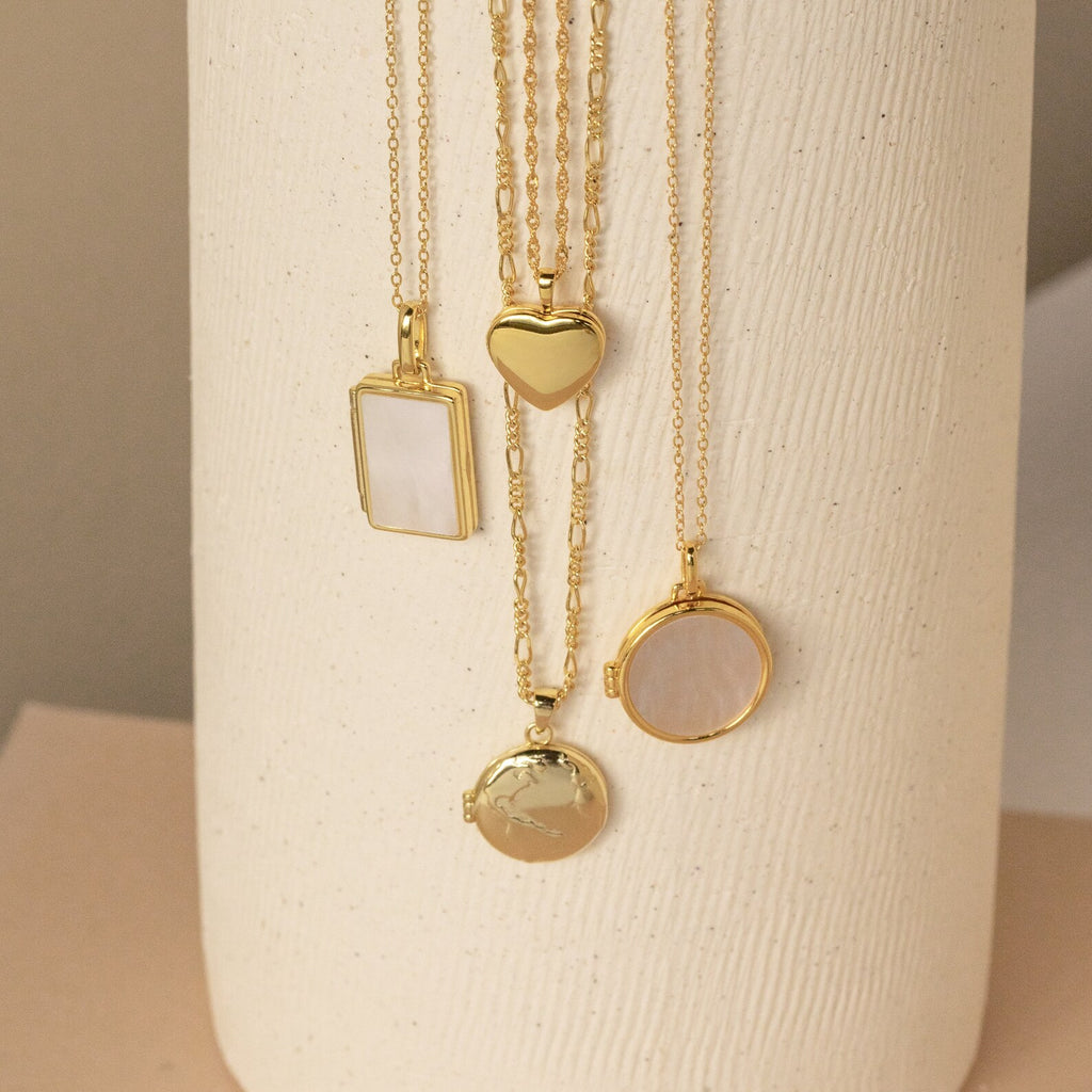 Four gold locket necklaces hang vertically on a cream textured vase—featuring rectangular, heart, and two round lockets, some with smooth polished finishes and others with glossy mother-of-pearl inlays.