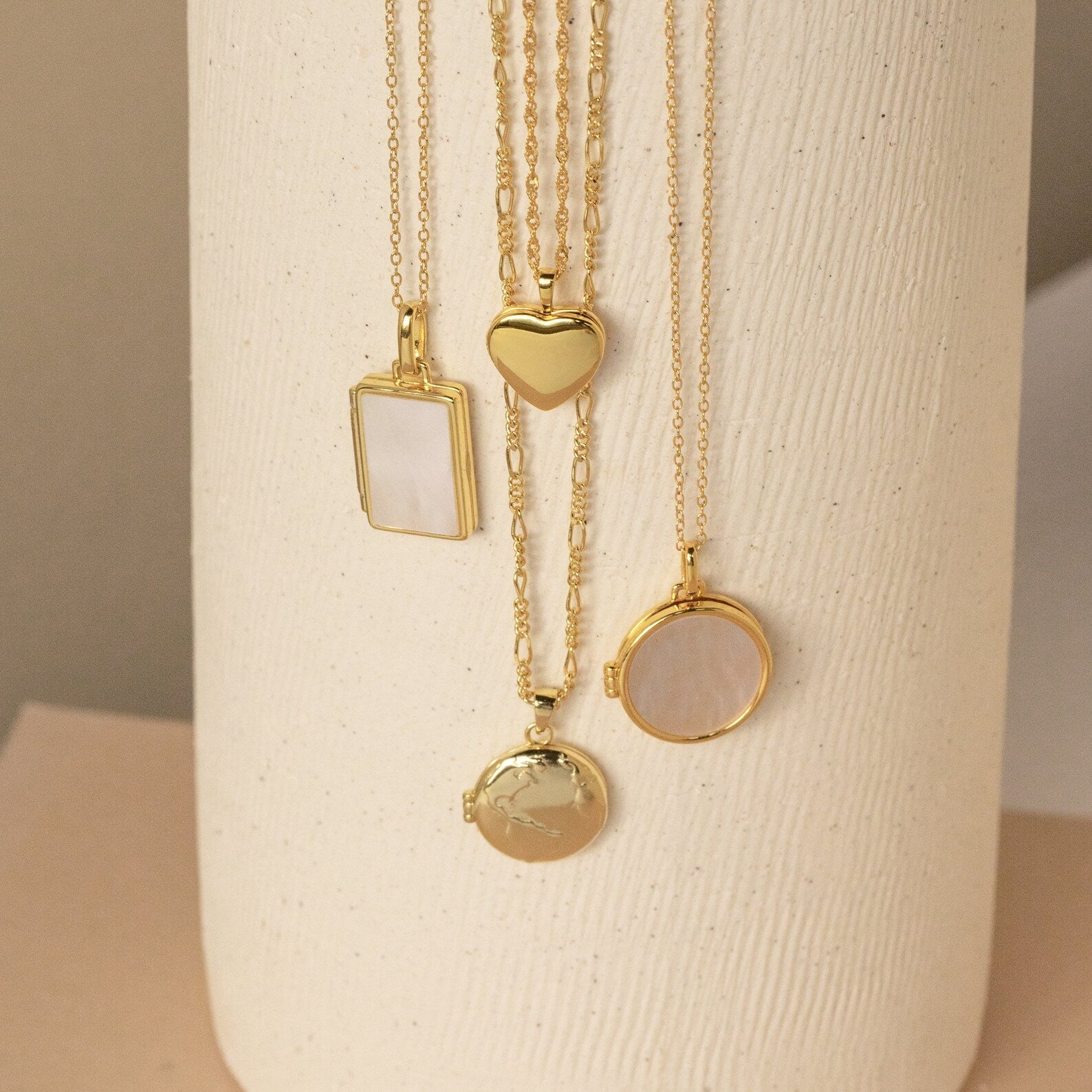 Four gold locket necklaces hang vertically on a cream textured vase—featuring rectangular, heart, and two round lockets, some with smooth polished finishes and others with glossy mother-of-pearl inlays.