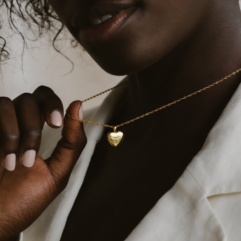 A woman in a white blazer holds an Initial Heart Locket Necklace close to her neck, highlighting elegant personalized jewelry.